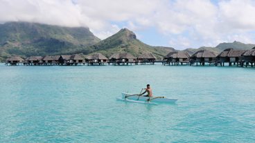 Man rows a boat in the ocean with bungalows in the background