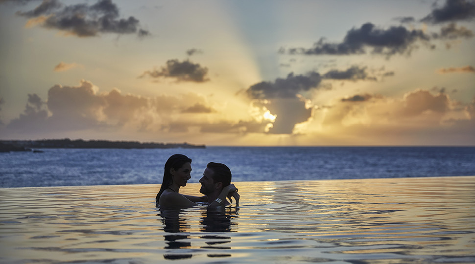 A man and woman embrace in a pool at sunset