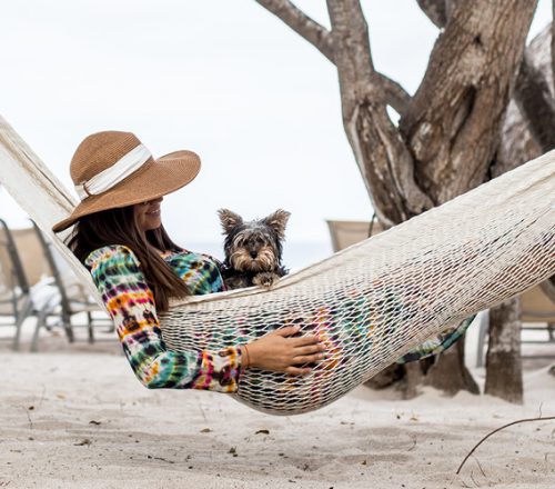 A woman in a straw hat sits in a hammock on the beach with a small dog in her lap