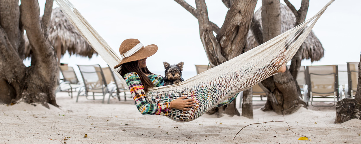 A woman in a straw hat sits in a hammock on the beach with a small dog in her lap