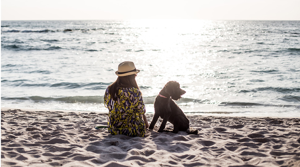A woman in a straw hat sits with a black dog on the beach