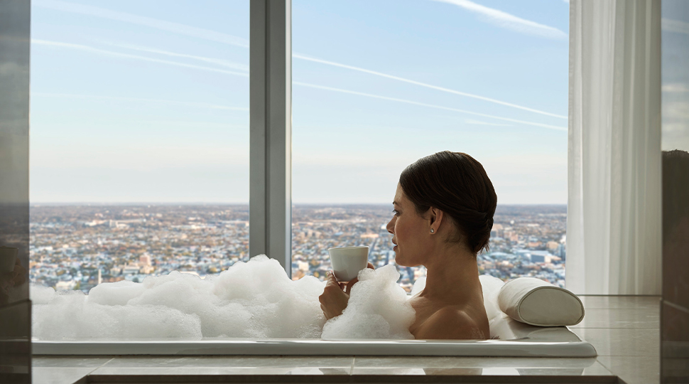 Woman with short dark hair sits in a bubble bath sipping a glass of champagne next to floor-to-ceiling views of a city skyline