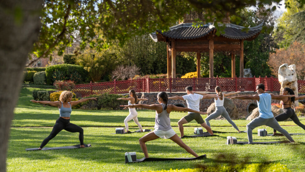 A yoga instructor teaching a class of six people outside near a wooden gazebo in a garden