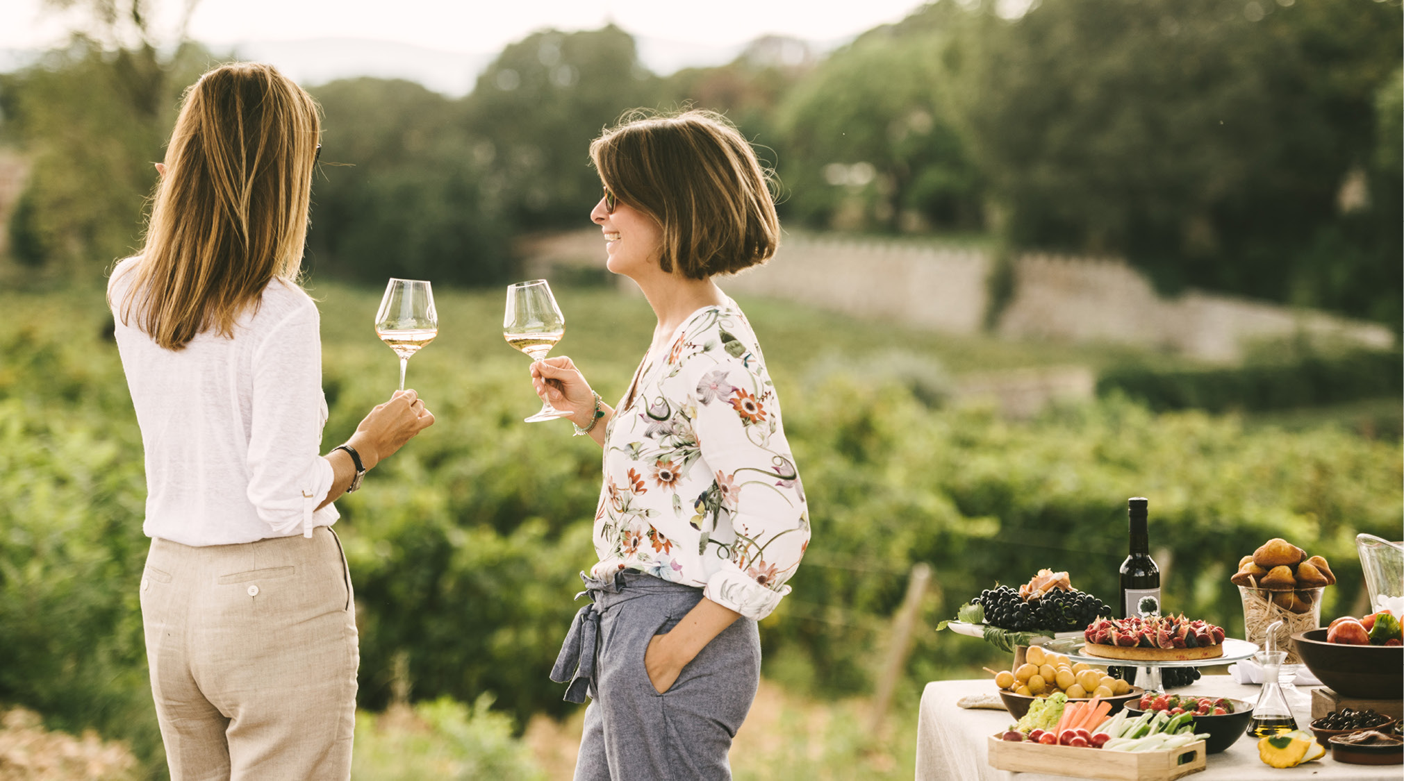 Two women drink a glass of white wine while standing among vineyards and private picnic