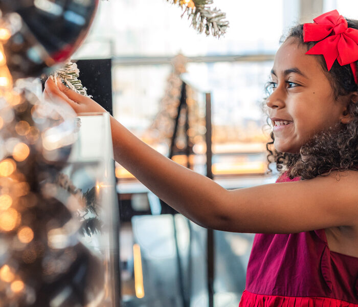 A young girl in a red dress touches the branch of a festive tree