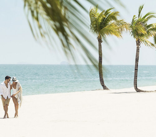 Two people walking arm-in-arm on a white-sand beach passing palm trees
