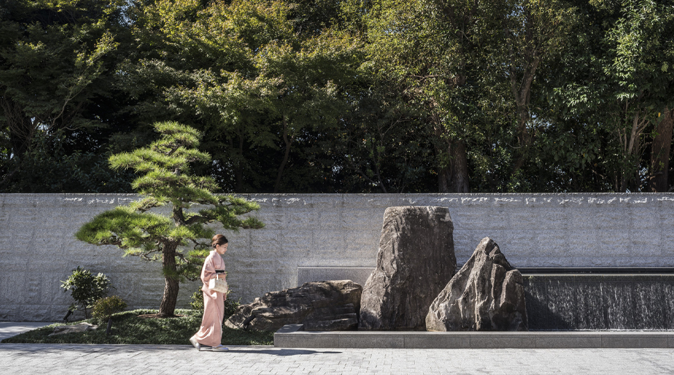 A geisha walks through a meditation garden featuring small trees, sand and stones