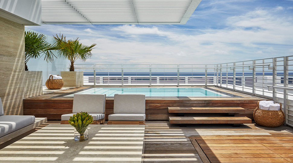 Outdoor deck featuring a square-shaped pool, two white lounge chairs, small palm trees and a view of the ocean in the background