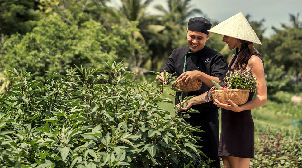 Man wearing a black chef's uniform helps a woman wearing a black dress and sunhat pick produce from a garden