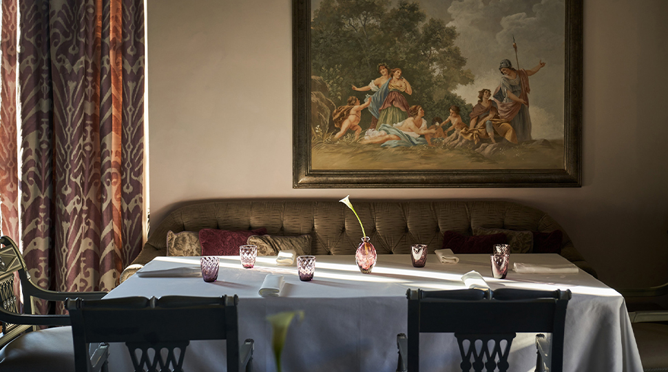 A white-linen topped table with a tufted banquette and four chairs sits beneath a Renaissance-style painting and red-a-gold curtains