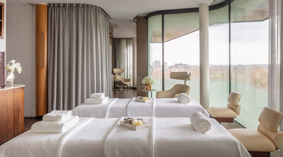 Two spa treatment beds with all-white linens and towels sit in the middle of a room next to floor-to-ceiling windows