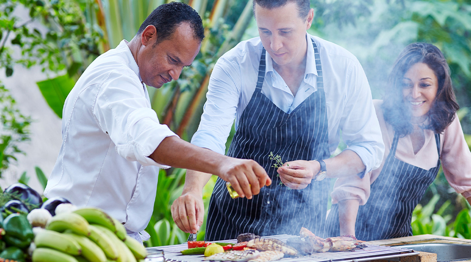 Three people stand over a grill seasoning vegetables