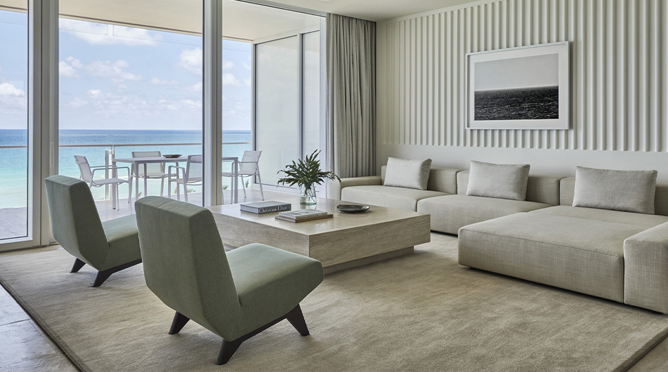 A light-gray sectional, two armless chairs and a large rectangular marble coffee table are all set next to floor-to-ceiling windows in a living room