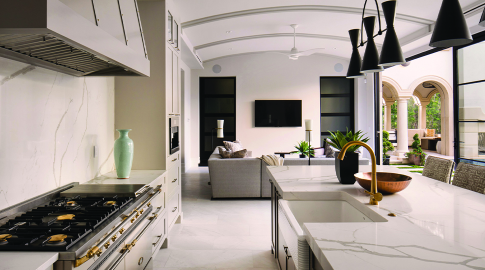 Large open-concept kitchen and living area with white marble countertops, industrial stove, black ceiling lamps, grey sectional and curved ceilings