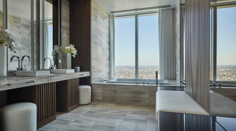 A spacious modern bathroom with double sinks, large mirror, a cushioned bench and a tiled bathtub overlooking a city view