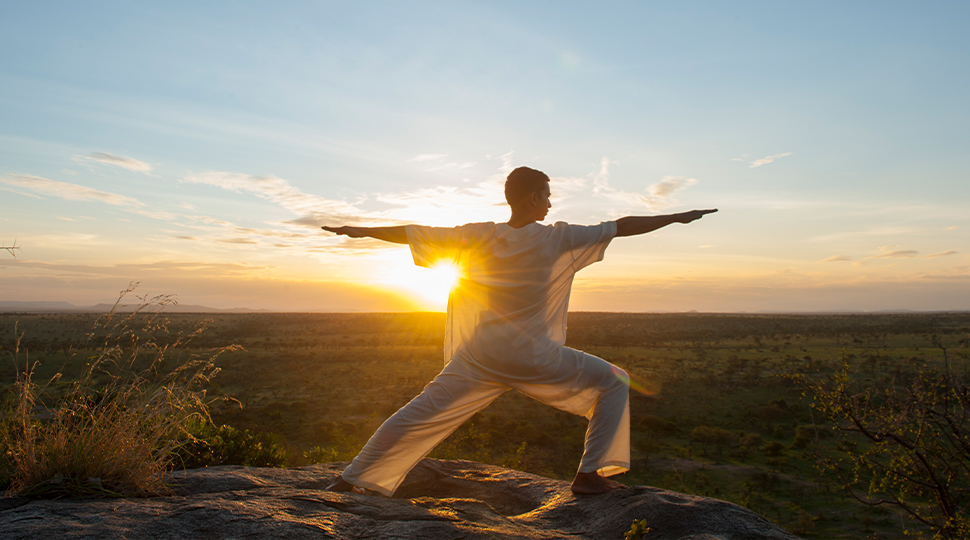 Person wearing all-white clothing stands in warrior pose at the top of a mountain with the sun setting in the background