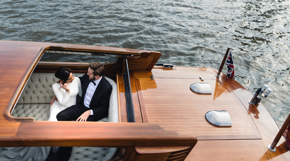 Couple dressed in formal wear sits close together in a wooden boat as it sails down the Thames River in London