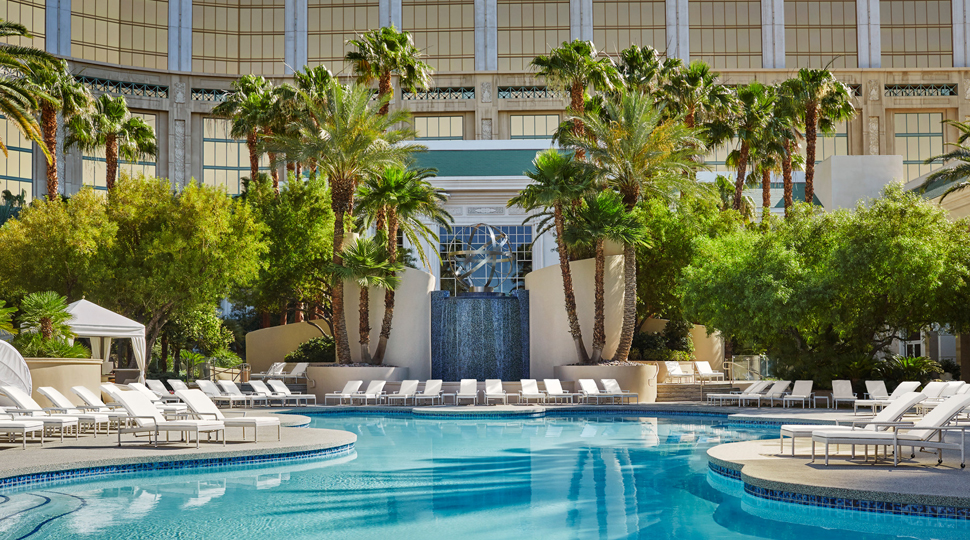 Outdoor pool surrounded by lounge chairs and trees, with a tiled waterfall in the background