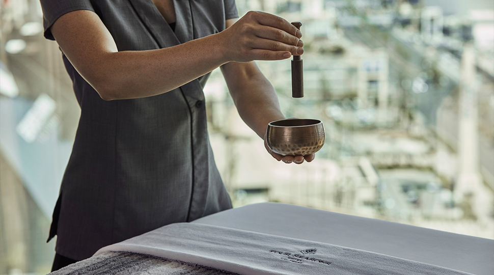 Spa attendant holding a small singing bowl and mallet over a treatment bed