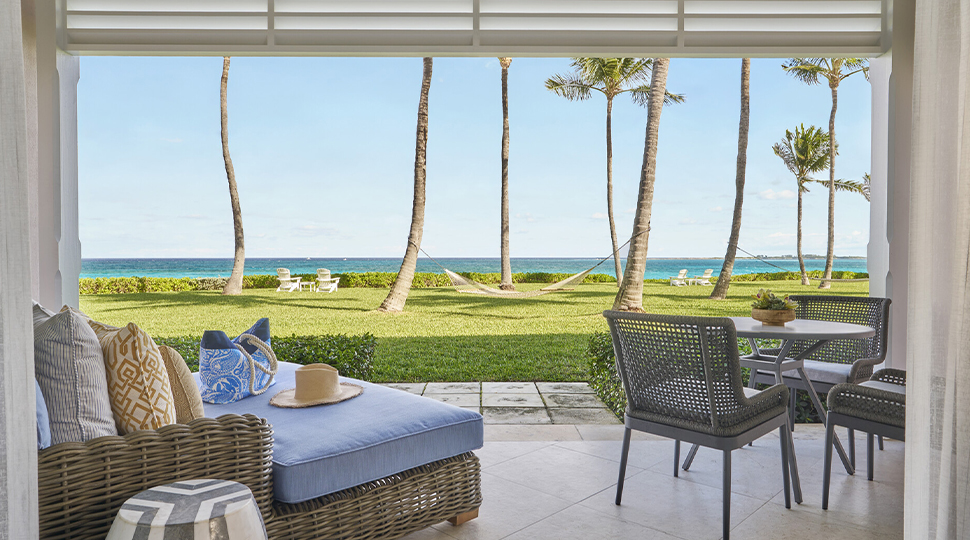 Patio of a guest room furnished with blue and rattan lounge furniture and a dining table and chairs overlooking a lawn with the ocean in the distance