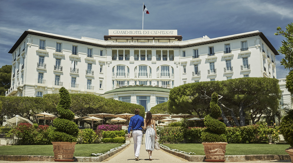 Couple walking arm in arm up to a white hotel building surrounded by green landscaped grounds