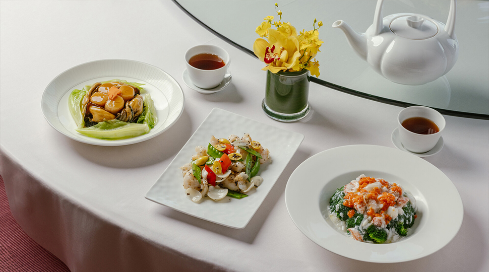 Three plates of food set on a white table along with two tea cups and a small vase of yellow flowers