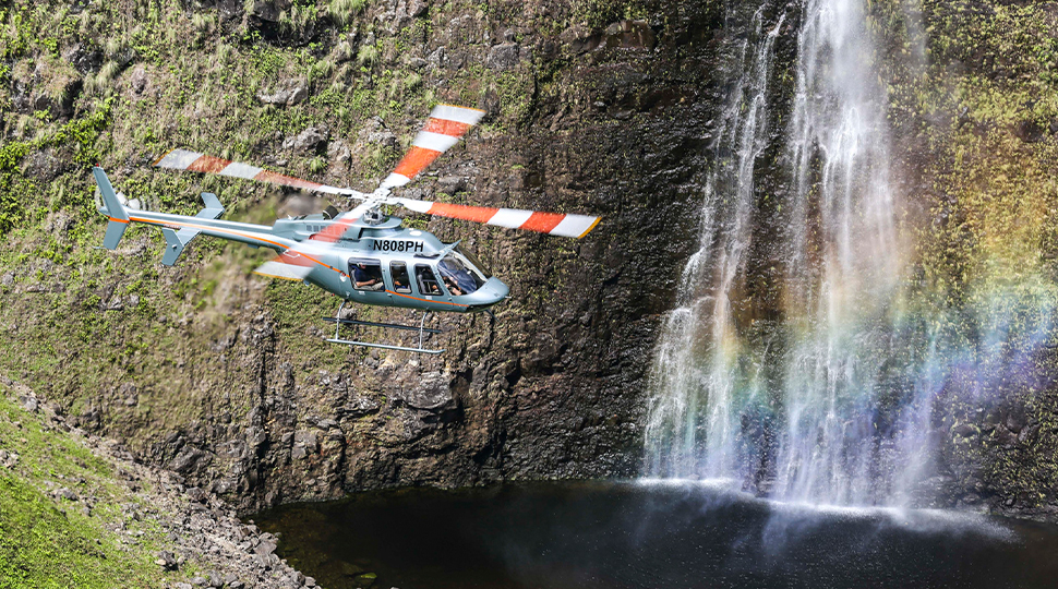 Helicopter flies in front of waterfall in Hawaii, with a rainbow seen in the mist