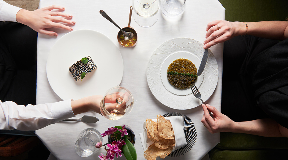 Overhead view of one person's hands and arms cutting into a dish on a white plate and another person's hands and arms across the table raising a glass