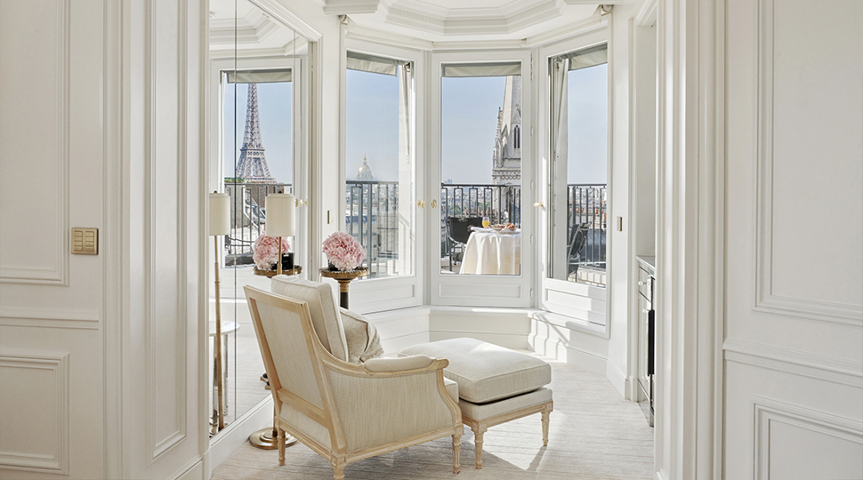 A wall of windows surrounded by white walls and floors look out onto the Eiffel Tower, with a white lounge chair and ottoman set beneath the windows