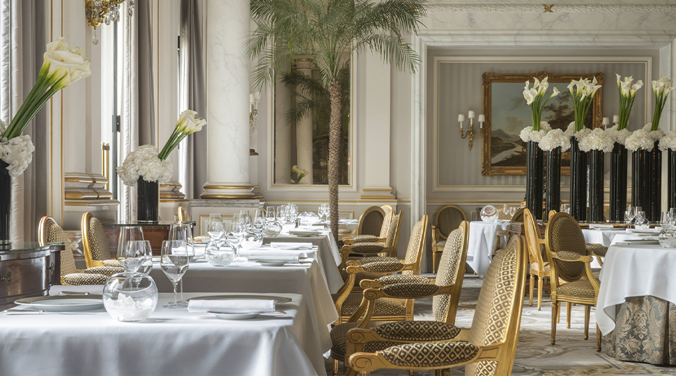 Interior of restaurant with white-clothed tables, gold-and-beige chairs and large white flowers in tall vases