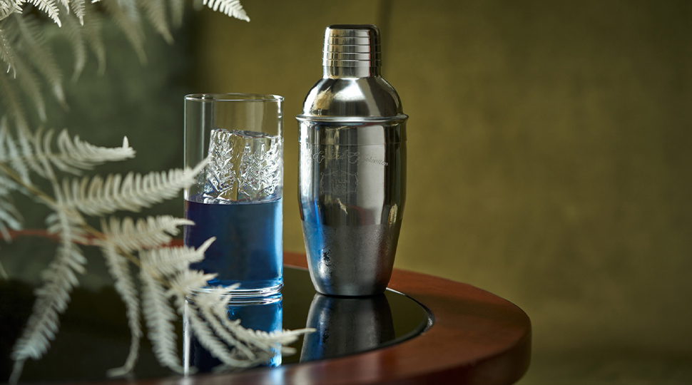 Silver cocktail shaker and a highball glass half-filled with a blue beverage sit on a cocktail table next to white tree branches