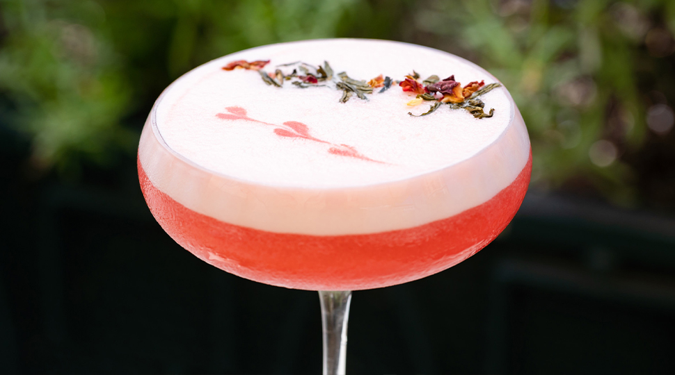 Rose-colored cocktail in a coupe glass topped with sprigs of flowers