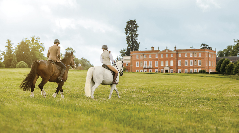 Two people riding horses, one brown horse and one white horse, on a grassy field with an English manor hous in the distance