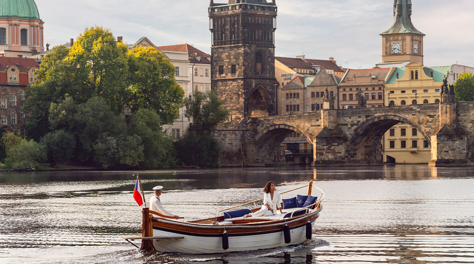 Two people ride in a small wooden boat along the river in Prague approaching Charles Bridge