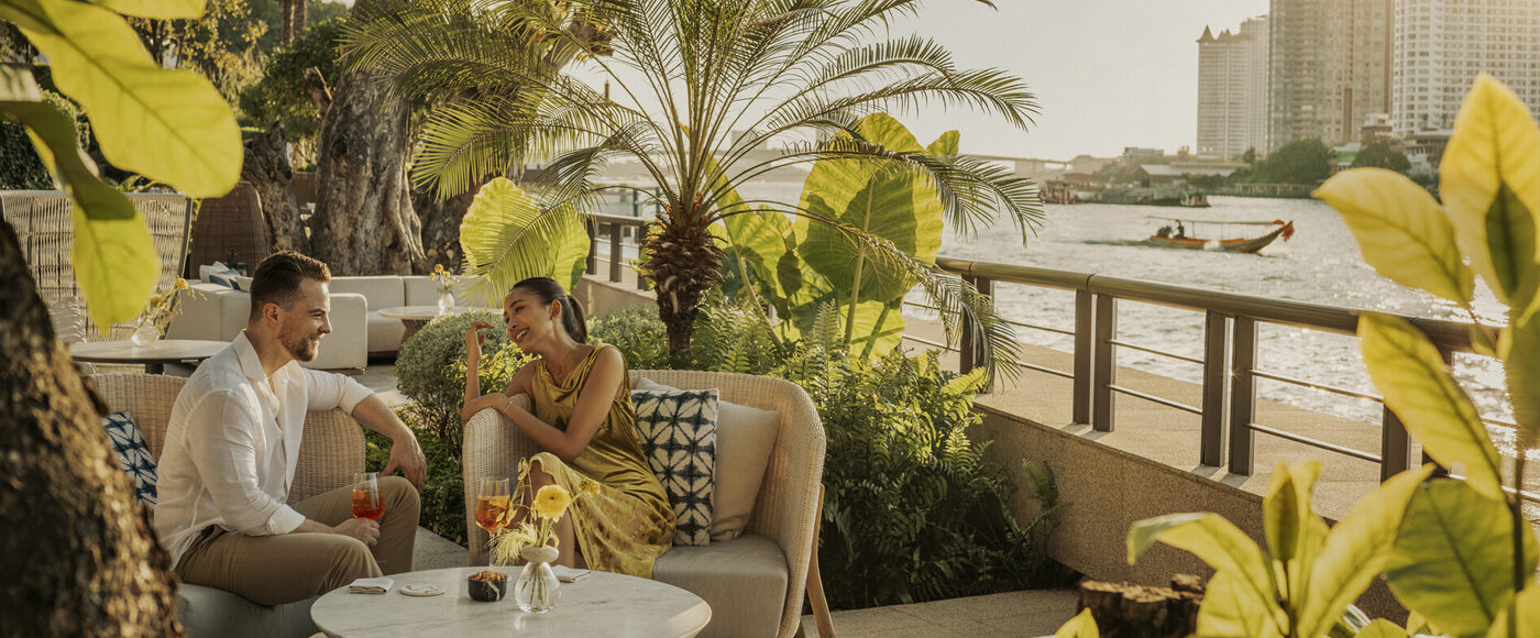 A man and a woman sit on rattan lounge chairs on an outdoor patio surrounded by large green foliage