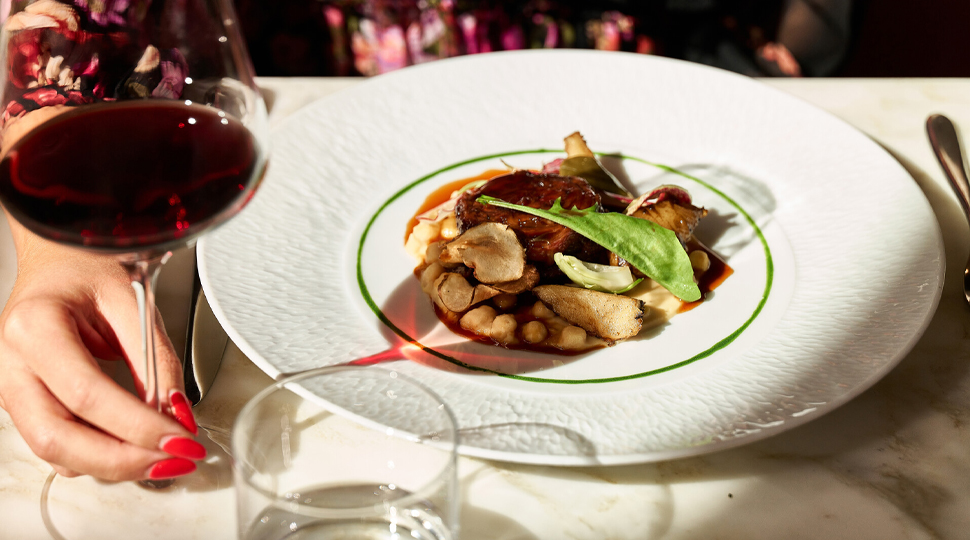 Braised lamb served on a white plate next to a glass of wine