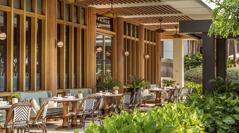 A row of tables set on an open-air restaurant terrace with large wood-framed widows on one side and lush greenery on the other