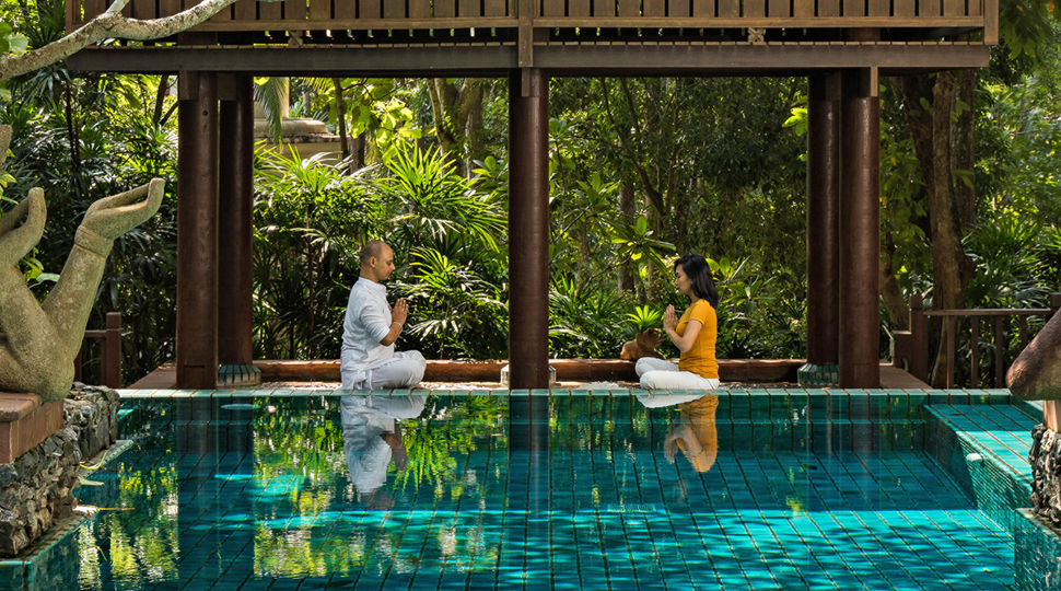 Two people sit cross-legged on the floor facing each other, both with their hands clasped together and eyes closed, on the ledge of a pool with a lush green jungle in the background