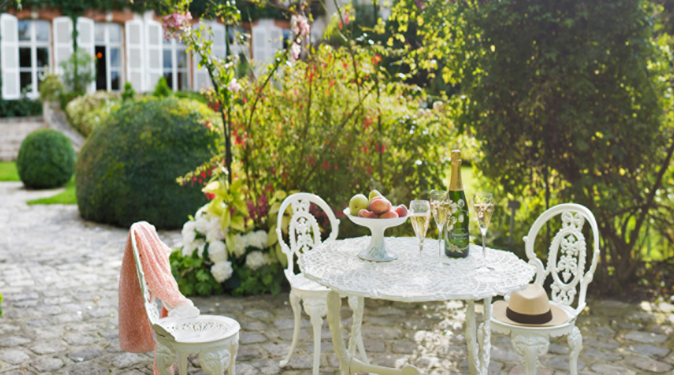 White wrought-iron bistro table with three chairs and a bottle of champagne, two glasses and a large bowl of fruit is set within a garden on a cobblestone patio