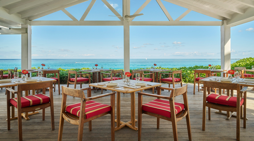 Outdoor, covered dining area with wood rectangular tables, chairs with red-and-white-striped cushions, all overlooking the ocean in the distance