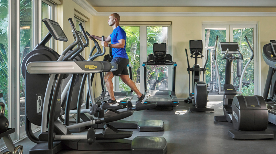 Person wearing blue t-shirt and black shorts running on a treadmill in a hotel gym with large windows