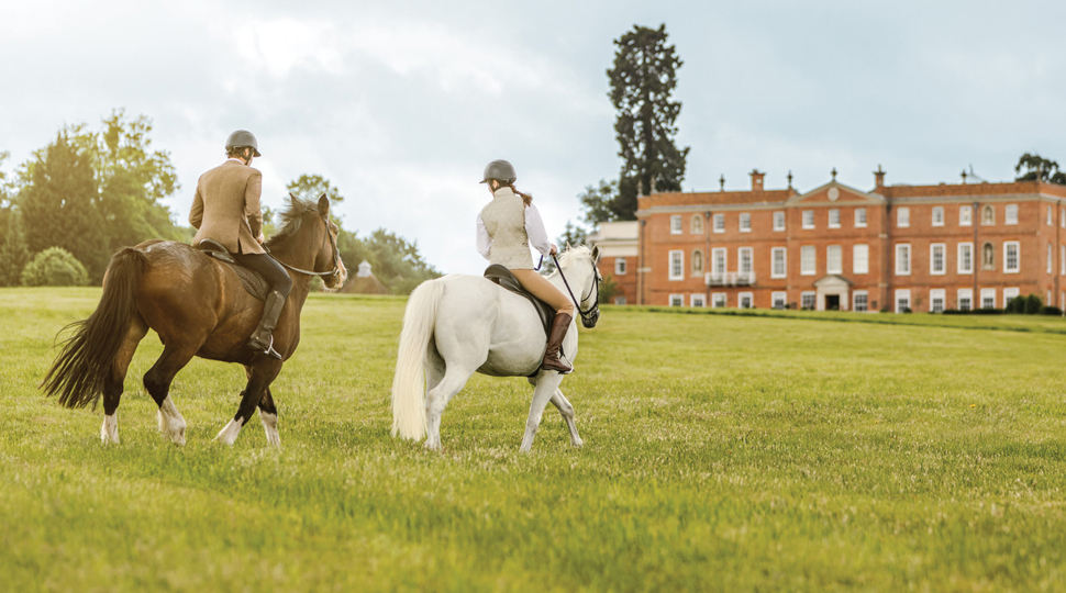 Two people ride horseback through the grounds of an English country estate