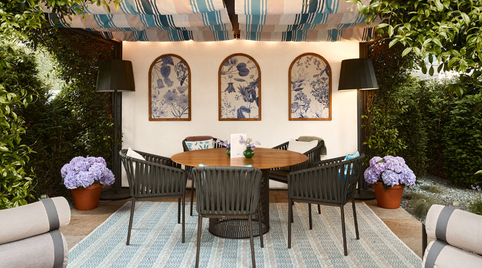 A round wooden table and five black chairs sit beneath a canopy at an outdoor terrace