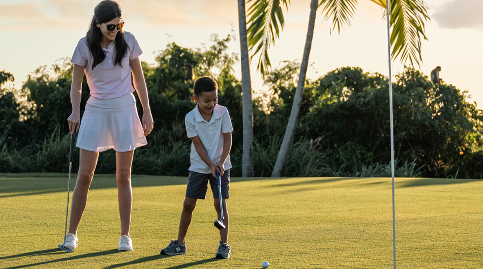 A parent watches their child putt a ball into the hole one a golf green