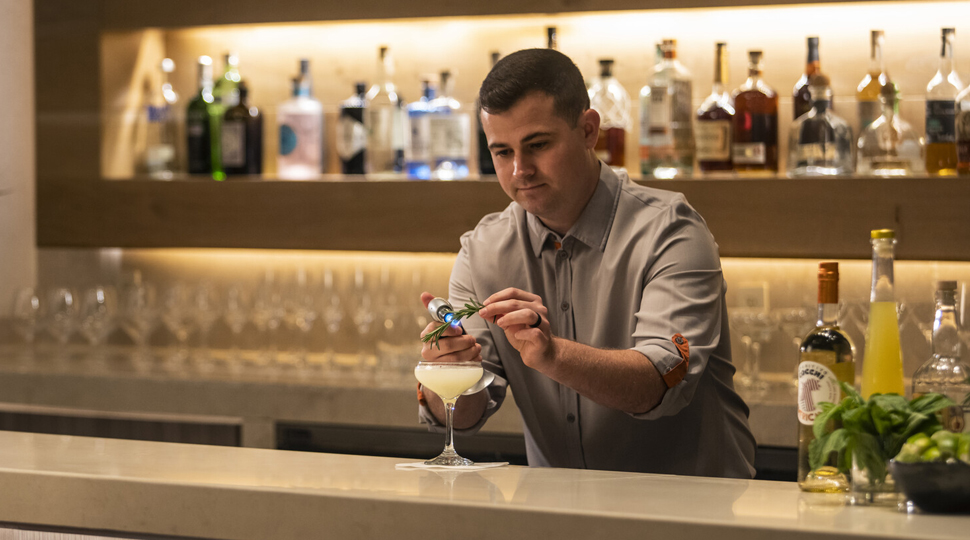 Bartender wearing light-blue, button-down shirt and light-grey vest puts a garnish on a cocktail