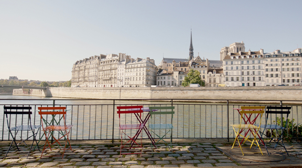 Three pairs of colourful bistro chairs line the banks of the Siene river