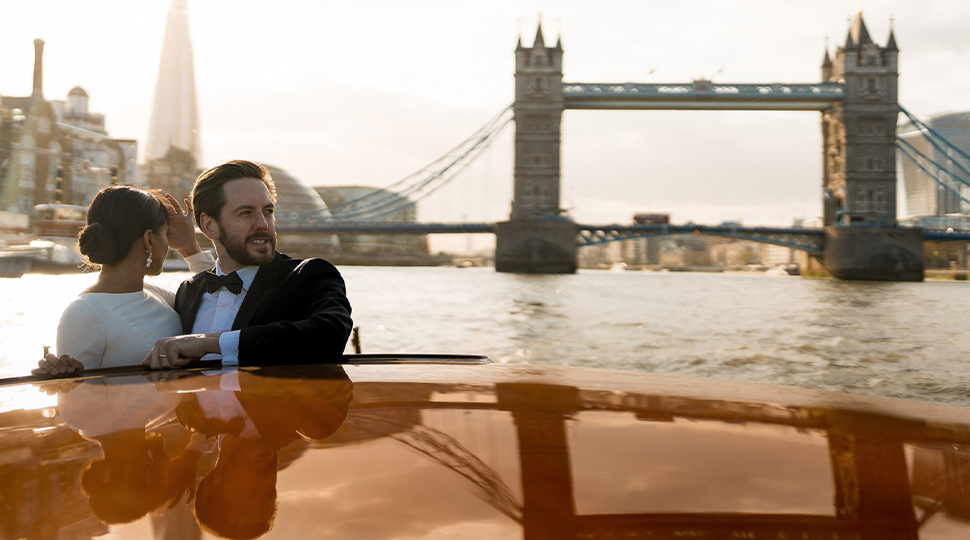 A couple dressed in formalwear peeks out of a small boat as they float by the Tower Bridge