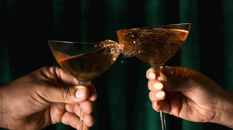 Two hands each holding coupe glasses filled with a beverage toast each other in front of a dark green backdrop