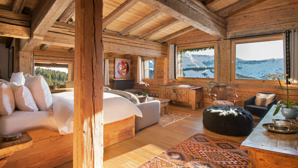 Bedroom of a mountain-top suite with wood-beamed ceilings, wooden posts a bed with white linens a sitting area and windows overlooking snow-covered mountains