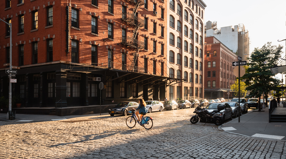 A person riding a bicycle in front of of a brick building with cars lining the street.